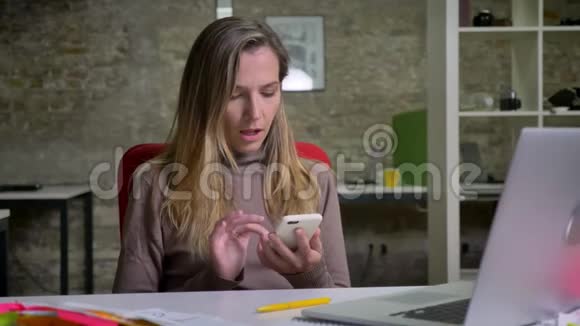现代女性员工在手机上浏览的特写镜头让她抓狂坐在笔记本电脑旁大笑视频的预览图
