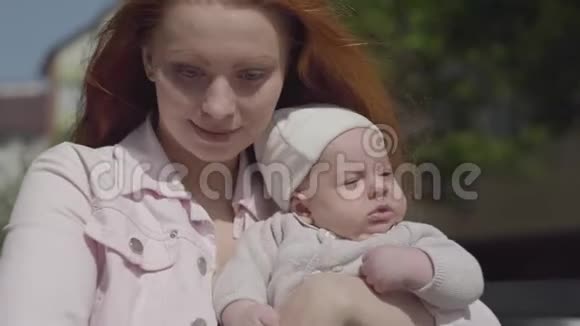 在春天阳光明媚的阳光下一位年轻漂亮的红发妈妈抱着可爱的宝宝和他说话视频的预览图