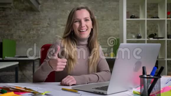 坐在笔记本电脑前的漂亮女办公室工作人员的特写肖像微笑着竖起大拇指视频的预览图