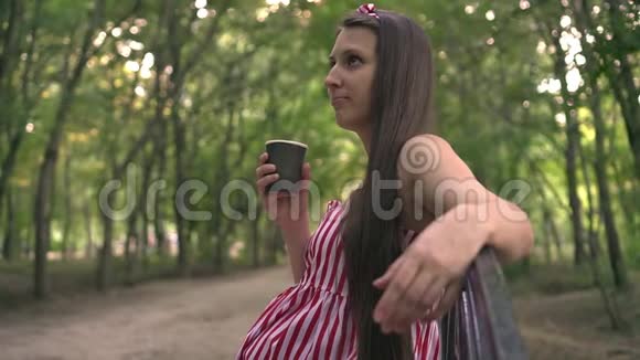 公园里绿树成荫的怀孕女孩一个穿条纹连衣裙的女孩坐在长凳上喝咖啡视频的预览图