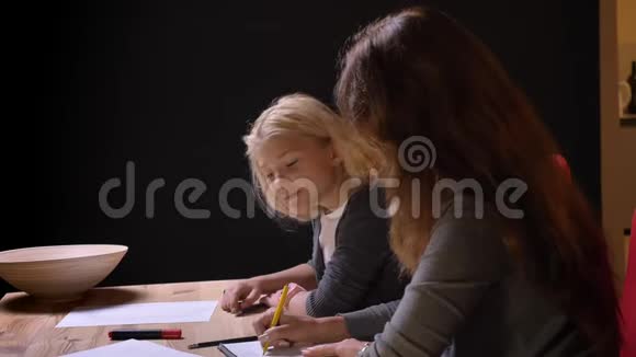 一位年轻的白种人母亲在舒适的室内家庭帮助她的小女孩做家庭作业拍摄特写镜头视频的预览图