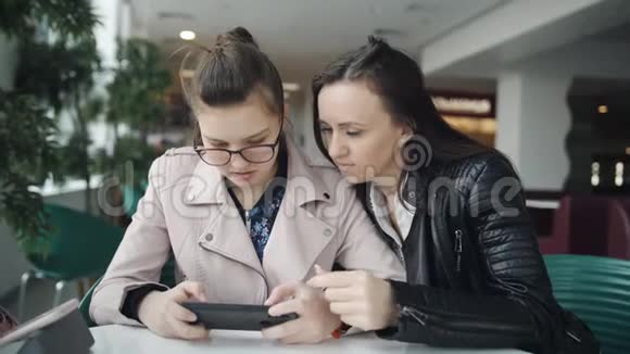 年轻的母亲和十几岁的女儿看着智能手机期待着咖啡馆的订单美女少女视频的预览图
