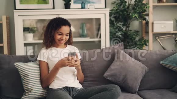 美丽的非裔美国女孩正在使用智能手机并在家里舒适的沙发上笑着放松现代技术视频的预览图