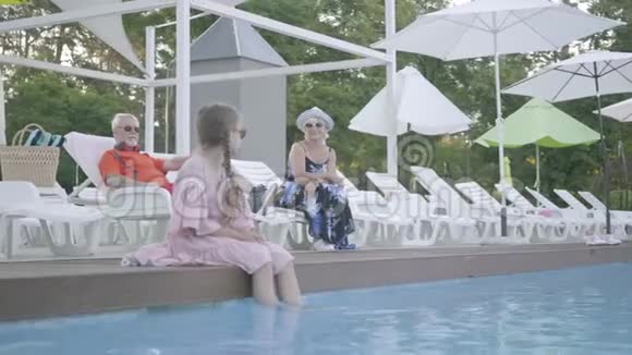 一对老夫妇躺在背景上靠近游泳池的日光浴床上坐在池边的小可爱视频的预览图