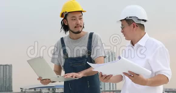 亚洲工程师站在屋顶上年轻的工程师用笔记本与年长的人讨论蓝图视频的预览图