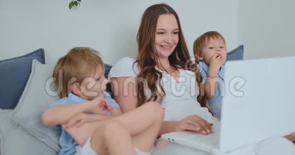 年轻漂亮的妈妈和两个孩子坐在沙发上看着笔记本电脑屏幕上网视频的预览图