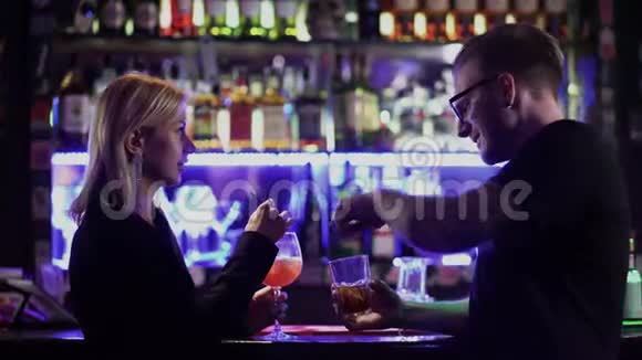 一对漂亮的夫妇在酒吧柜台喝鸡尾酒戴眼镜的男人和金发女人在迪斯科舞厅聊天玩得开心视频的预览图