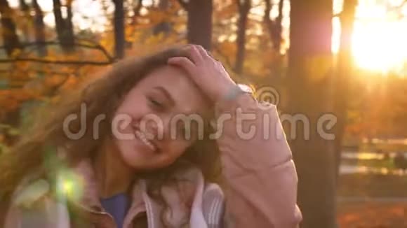 年轻的白种人卷发女子在闪亮的秋日公园背景下高兴地观看镜头的肖像视频的预览图
