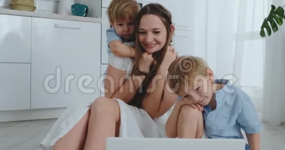 妈妈和两个孩子玩坐在地板上拥抱和玩一个幸福的家庭看完手提电脑后的游戏视频的预览图