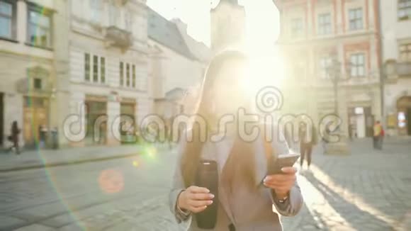 女人手里拿着一个保温杯在夕阳下用智能手机沿着一条老街走着通信社交网络视频的预览图
