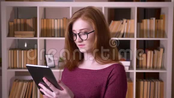 在大学图书馆里用平板电脑拍摄的戴眼镜的年轻红发美女白种人的特写照片视频的预览图