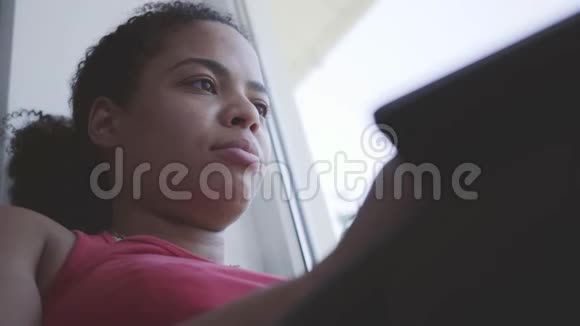 可爱的年轻非洲裔美国妇女坐在窗台上在她的笔记本电脑上打字快乐的女孩在家放松休息日视频的预览图