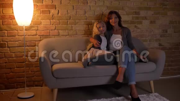 年轻的白种人女性和她的小漂亮女儿坐在沙发上拥抱看电视的特写镜头视频的预览图