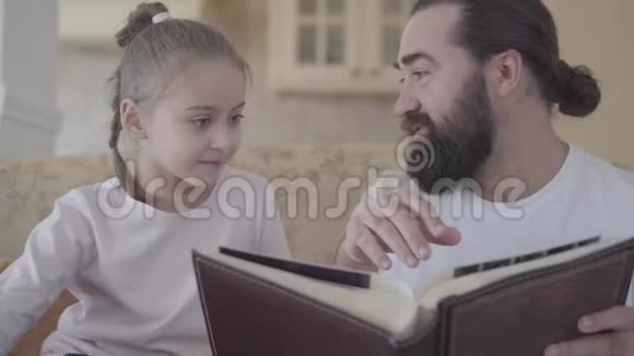 大胡子坐在沙发上向他的小女儿展示书幸福家庭的观念小女孩视频的预览图