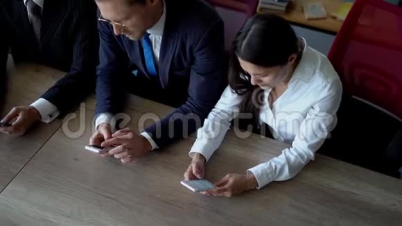 两个商人和女商人坐在办公室的桌子上一边浏览手机视频的预览图