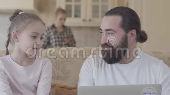 大胡子男人坐在沙发上和他的小女儿近距离交谈幸福家庭的观念小女孩分享视频的预览图