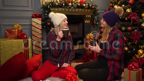 两个穿着红色格子衬衫的女孩坐在圣诞树下戴着温暖的白色和紫色的帽子谈论她们的事情视频的预览图