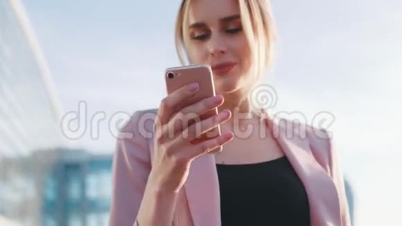 一个可爱的金发女人穿着粉红色夹克用手机在明亮的阳光下发短信外部视频的预览图