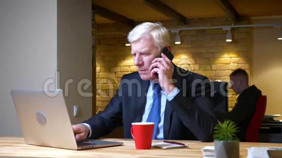 一个老白种人商人在办公室里用手提电脑打电话的特写镜头视频的预览图