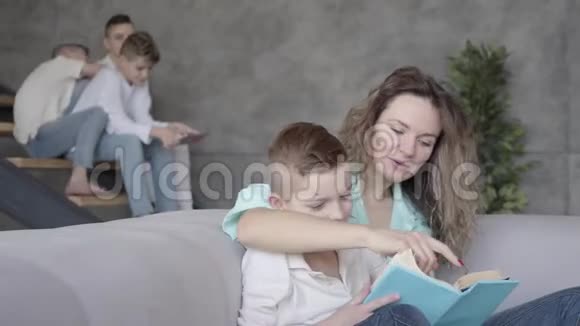 年轻漂亮的母亲给儿子看书的肖像而其余的孩子则在玩耍视频的预览图