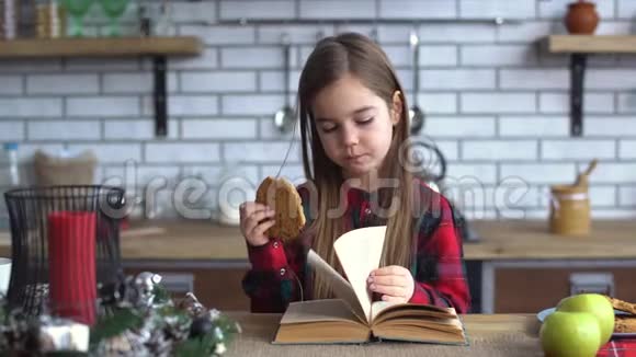 可爱的小女孩穿着格子衬衫留着长发在厨房的桌子上吃饼干翻阅一本书视频的预览图