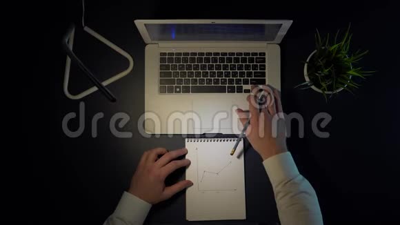 一个男人坐在桌子旁晚上坐在笔记本电脑前在笔记本上画一张收入图4k视频的预览图