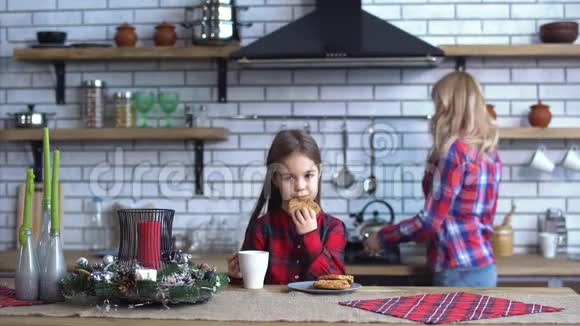 棕色长发的小女孩在厨房的桌子上吃着带茶的饼干年轻的母亲在煮咖啡视频的预览图