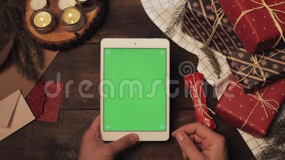 手的顶部视图滚动和粘贴在垂直绿色屏幕上的全新白色平板电脑在木制桌子上与圣诞节视频的预览图