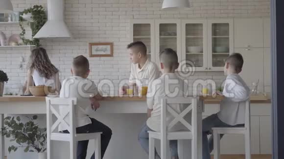 年轻漂亮的母亲为她的四个穿着白衬衫的十几岁的孩子烤馅饼等待着坐在桌子旁的食物视频的预览图