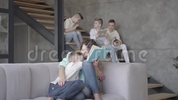 年轻漂亮的母亲在给儿子读书而其余的孩子则在后台互相玩耍视频的预览图