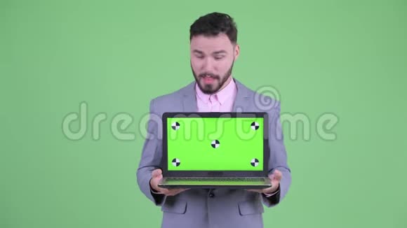 快乐的年轻大胡子商人一边说话一边展示笔记本电脑视频的预览图