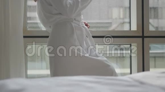 穿着白色浴袍的年轻女子站在酒店公寓的窗户旁手里拿着智能手机和一杯咖啡关门视频的预览图