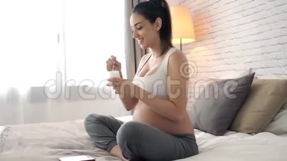 美丽的年轻孕妇在家坐在床上吃酸奶视频的预览图