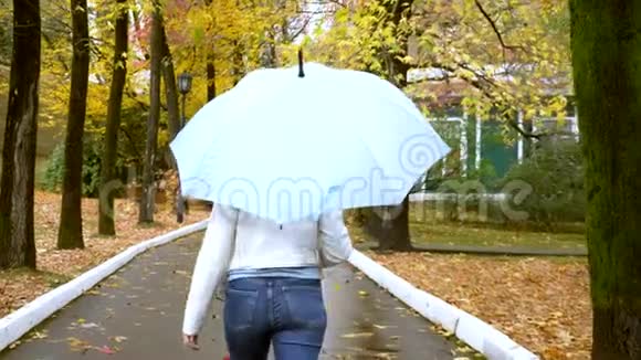 穿着白色皮夹克和牛仔裤的瘦女人在秋天的公园里带着蓝色的雨伞在雨中散步视频的预览图