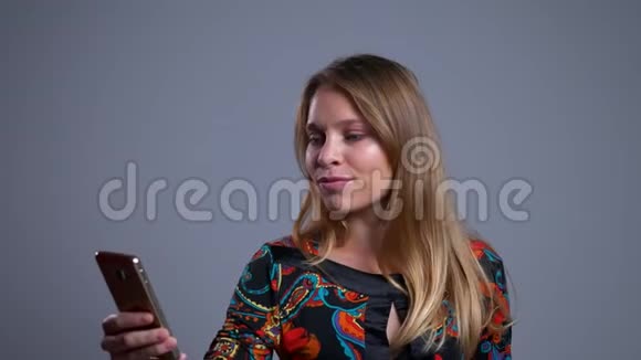 一幅美丽的年轻白种人女子的特写肖像正带着积极的微笑在电话中观看照片视频的预览图