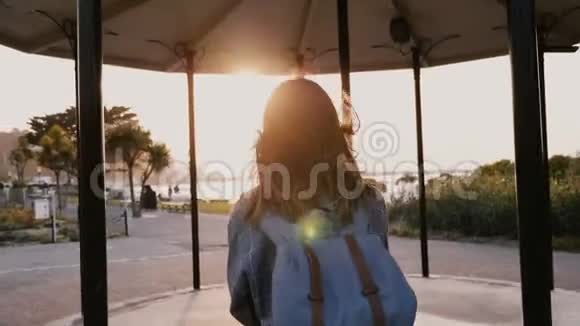 镜头跟随快乐休闲的年轻女子背着背包头发随风飘散沿着夏日夕阳西下的海滨公园漫步视频的预览图