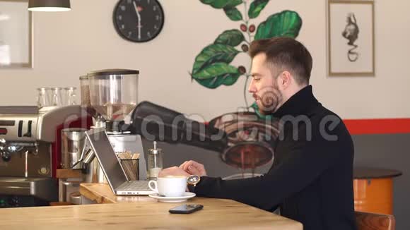 一位留胡子的年轻商人在咖啡店的肖像他在笔记本电脑上工作视频的预览图