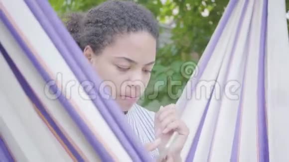 年轻的非洲裔美国妇女坐在吊床上在花园里放松用手机发短信视频的预览图