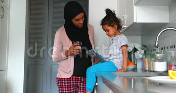 年轻的母亲戴着头巾在厨房的女儿旁边喝着一杯水视频的预览图