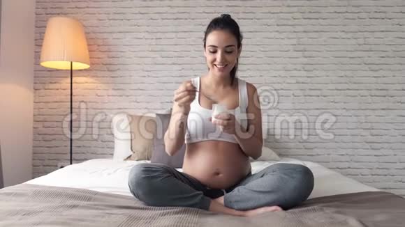 美丽的年轻孕妇在家坐在床上吃酸奶视频的预览图