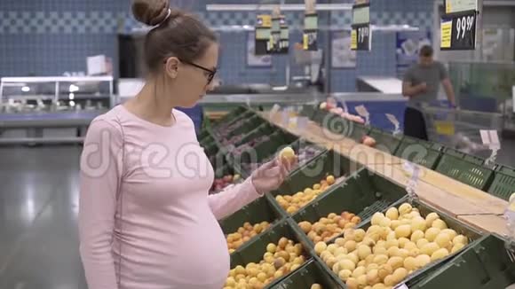 在超市里戴眼镜的年轻孕妇选择新鲜的有机水果女人在购物时选择水果视频的预览图
