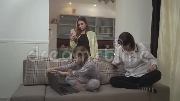 两个姐姐在手机上发短信小女孩坐在房间里的沙发上用笔记本电脑打字小可爱视频的预览图