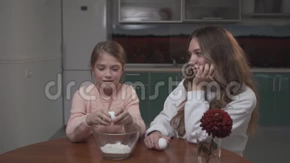 女孩和姐姐坐在厨房的桌子旁在碗里打碎了一个鸡蛋视频的预览图