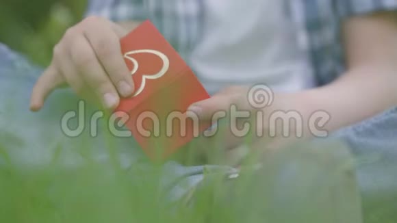 男孩拿着一个红色的小盒子打开准备礼物给女孩坐在户外的草地上夏季视频的预览图