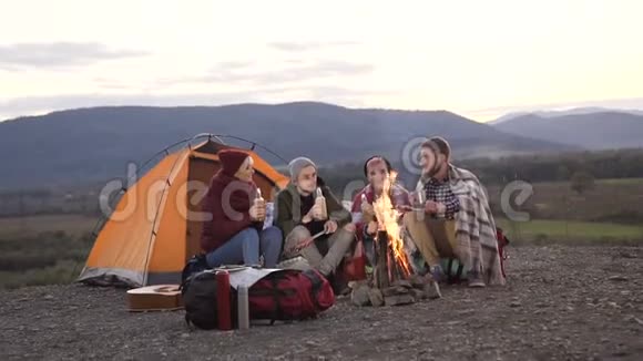 四个白种人的朋友在旅游帐篷附近休息篝火在山上炸香肠山脉视频的预览图