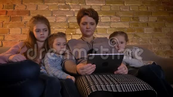 一幅白种人母亲和三个女儿的家庭画像她专注地看着平板电脑对舒适感兴趣视频的预览图
