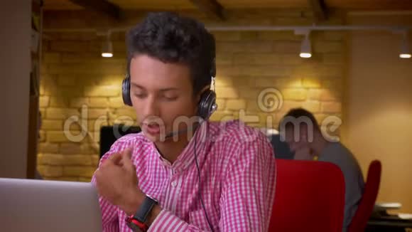 印度年轻商人戴着耳机用笔记本电脑在室内办公室打电话的特写镜头视频的预览图