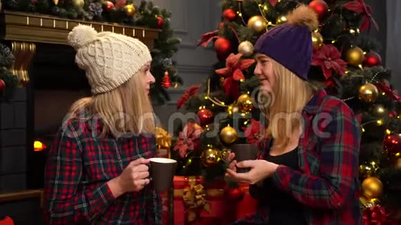 两个穿着红色格子衬衫的女孩坐在圣诞树下戴着温暖的白色和紫色的帽子谈论她们的事情视频的预览图