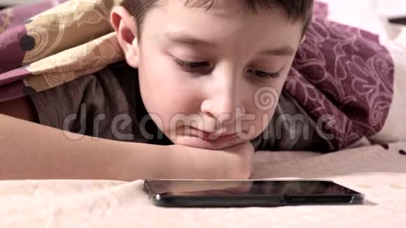 一个男孩躺在家里的床上在智能手机上放松微笑和观看视频视频的预览图