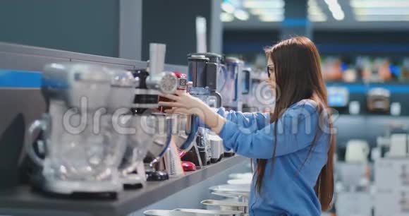 在电器商店里一个穿着衬衫的黑发女人通过观看和拿着设备来选择搅拌机购物视频的预览图